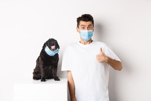 Covid-19, animals and quarantine concept. Image of funny young man and small dog in medical masks, owner showing thumb up in approval, praise something, white background.