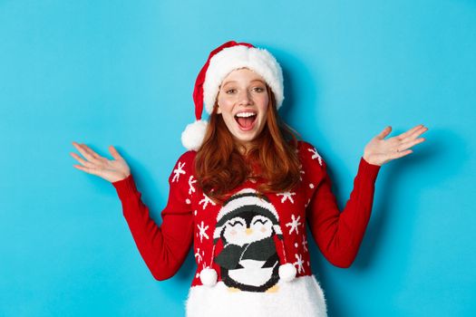 Happy holidays concept. Cheerful redhead girl in xmas sweater and santa hat, raising hands up and wishing merry Christmas, standing against blue background.
