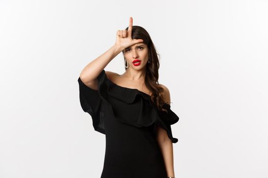 Fashion and beauty. Arrogant glamour woman showing loser sign on forehead, mocking lost person, standing in black dress over white background.