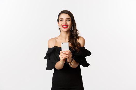 Attractive party girl taking photo on smartphone, make photograph on mobile phone camera, standing in black dress over white background.