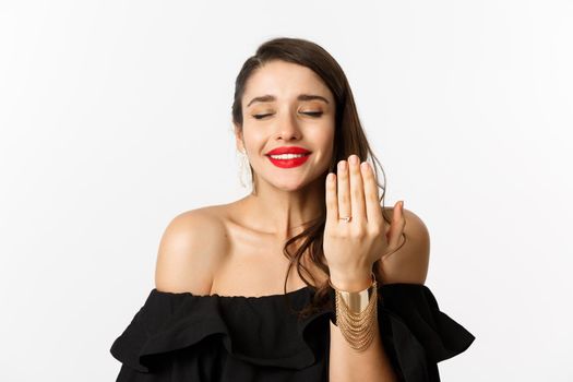 Happy young woman saying yes, become a bride, showing engagement ring on finger and smiling pleased, standing over white background.