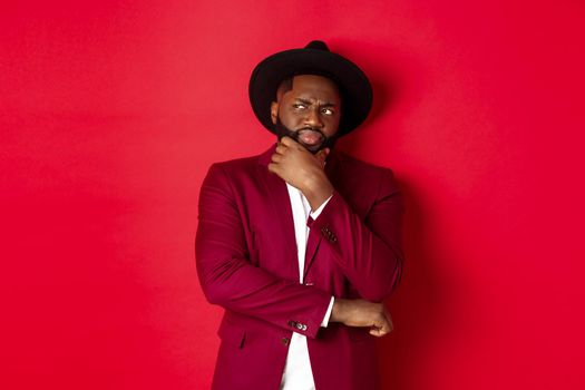 Fashion and party concept. Skeptical and doubtful african american man thinking, looking displeased at upper left corner, having doubts, red background.