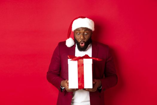 Christmas, New Year and shopping concept. Surprised Black man staring at xmas present, saying wow amazed, receiving holiday gift, red background.