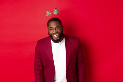 Merry Christmas. Handsome african american man in blazer and party headband, celebrating new year, smiling happy at camera, red background.