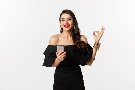 Online shopping concept. Attractive woman in trendy black dress, makeup, showing okay sign in approval and using mobile phone app, white background.