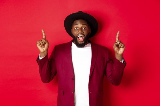 Winter holidays and shopping concept. Cheerful african american man in party outfit pointing fingers up, showing logo and smiling happy, red background.