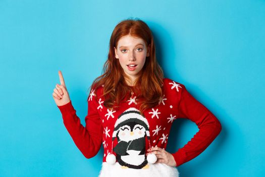 Winter holidays and Christmas Eve concept. Sassy redhead girl in xmas sweater, pointing upper left corner and staring at camera, hinting on promo offer, blue background.