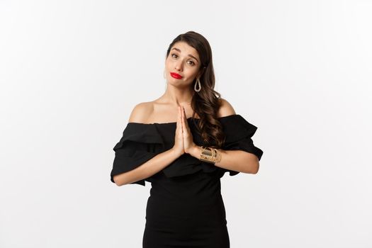 Fashion and beauty. Pretty stylish woman asking for help, begging for something, say please with hands pressed together, standing in black dress, white background.