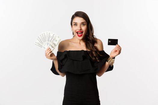 Fashion and shopping concept. Excited woman in black dress, showing credit card and dollars, smiling and staring at camera, white background.