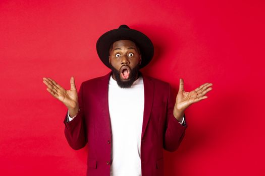 Fashion and party concept. Impressed Black man in classy outfit staring with complete disbelief at camera, gasping and looking surprised, standing over red background.