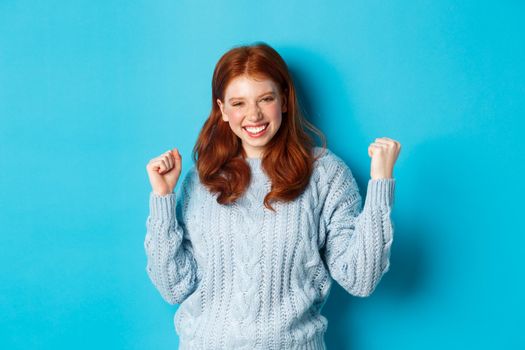 Satisfied redhead girl achieve goal and celebrating, making fist pump gesture and smiling delighted, triumphing of win, standing against blue background.