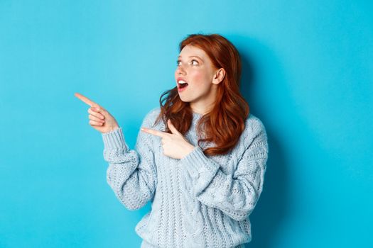 Winter holidays and people concept. Amazed redhead girl checking out promo offer, pointing and looking left at copy space, standing against blue background.