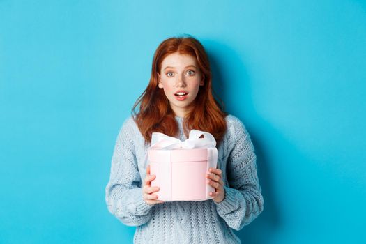 Surprised redhead girl receiving valentines gift, holding box with present and staring at camera amazed, wearing sweater, standing over blue background.