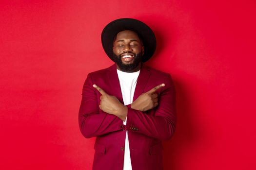 Happy young man dreaming and pointing fingers sideways, close eyes and smiling, showing two things, standing over red background.