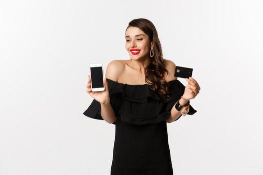 Fashion and online shopping concept. Happy young woman in black dress, showing credit card and mobile screen, standing over white background.