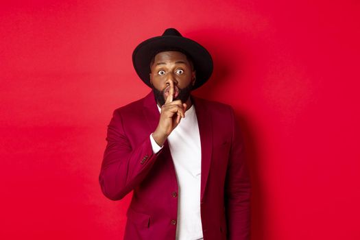 Fashion and party concept. African american man in classy hat hushing, asking keep secret, preparing surprise, standing over red background.