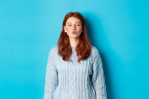 Cute redhead teen girl waiting for kiss, pucker lips and close eyes, standing in sweater against blue background.