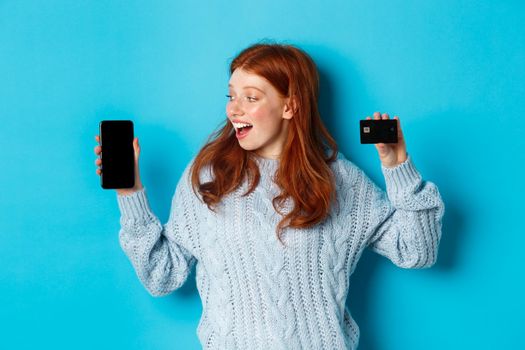 Excited redhead girl showing mobile phone screen and credit card, demonstrating online store or application, standing over blue background.