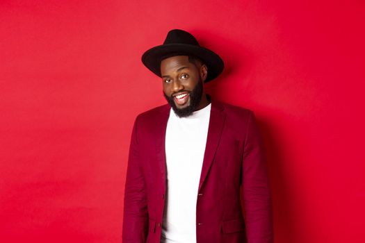 Christmas shopping and people concept. Handsome bearded african american guy looking at camera, smiling confident, wearing party clothes, red background.