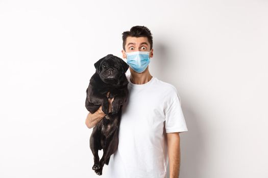 Covid-19, animals and quarantine concept. Funny young man in medical mask holding cute black pug dog, standing over white background.