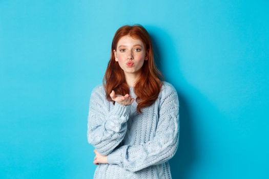 Lovely teen girl in sweater blowing air kiss, pucker lips and staring at camera, standing against blue background.
