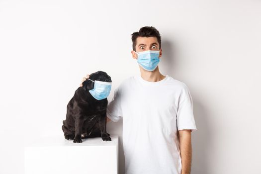 Covid-19, animals and quarantine concept. Image of funny young man and pug in medical masks, staring at camera, standing over white background.