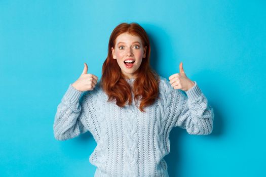 Amazed redhead girl in sweater, showing thumbs up and praising product, looking at camera amazed, standing over blue background. Copy space