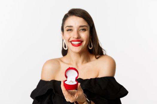 Fashion and beauty concept. Close-up of gorgeous brunette woman with red lips, black dress, showing engagement ring in box and smiling, getting married, standing over white background.