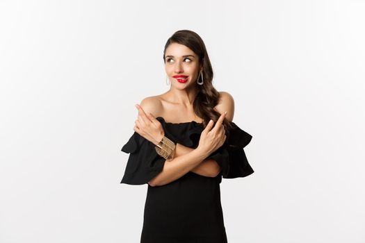 Fashion and beauty. Beautiful glamour woman in black dress, making choice, biting lip from temptation and pointing sideways, standing over white background.