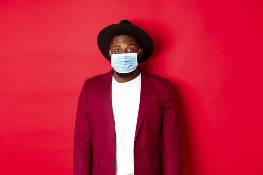 Covid-19 and fashion concept. Handsome african american man in stylish blazer and hat, wearing medical mask from coronavirus, standing over red background.