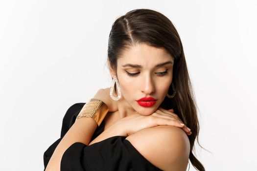 Fashion and beauty concept. Close-up of sensual woman in elegant earrings and black dress, wearing makeup with red lips, looking down tenderly, standing over white background.