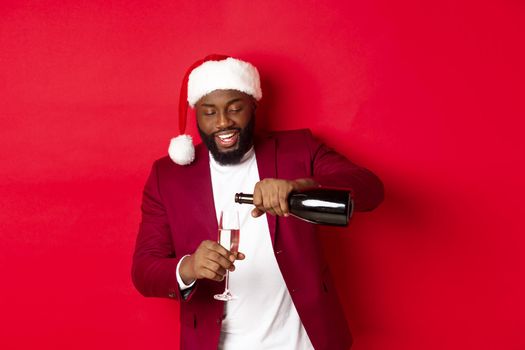 Christmas, party and holidays concept. Handsome Black man in santa hat pouring glass of champagne and smiling, celebrating New Year, red background.