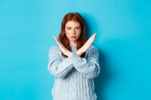 Serious redhead girl looking confident, showing cross gesture to stop and forbid action, standing over blue background.