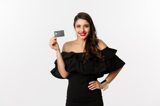 Beauty and shopping concept. Stylish pretty woman with red lips, black dress, smiling happy and showing credit card, standing over white background.