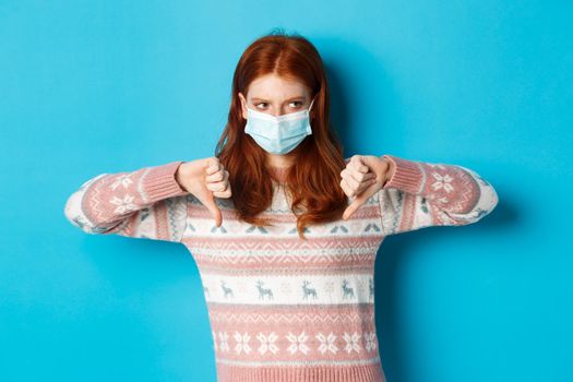 Winter, covid-19 and pandemic concept. Moody redhead girl in face mask showing thumbs-down, disapprove and dislike, standing against blue background.