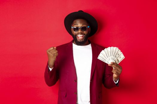 Cheerful african american guy in sunglasses, black hat and blazer, winning prize money, holding dollars and looking satisfied, standing over red background.