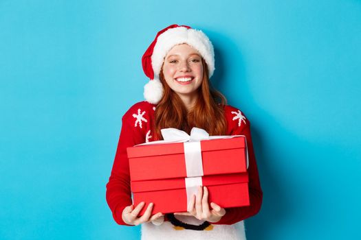Happy holidays and Christmas concept. Cheerful teen girl with red hair, wearing santa hat and holding xmas presents.