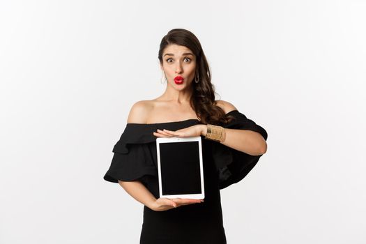 Fashion and shopping concept. Beautiful woman with red lipsticks, black dress, showing tablet screen and looking excited, standing over white background.