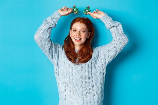 Winter holidays and Christmas sales concept. Beautiful redhead female model celebrating New Year, wearing funny party headband and sweater, smiling at camera.