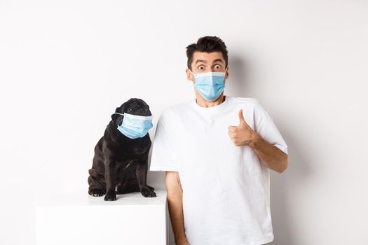 Covid-19, animals and quarantine concept. Image of funny young man and small dog in medical masks, owner showing thumb up in approval or like, white background.