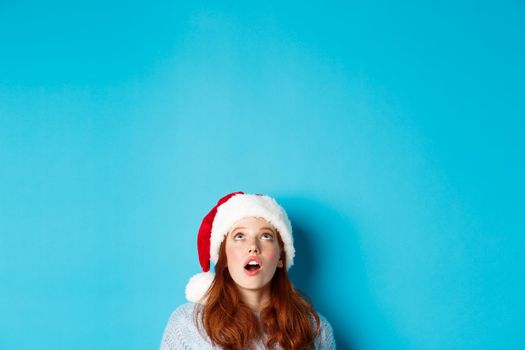 Winter holidays and Christmas eve concept. Head of pretty redhead girl in santa hat, appear from bottom and looking up at logo, seeing promo offer amazed, blue background.