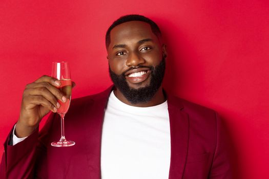 Close-up of handsome smiling Black man saying toast, raising glass of champagne for merry christmas and happy new year, celebrating against red background.