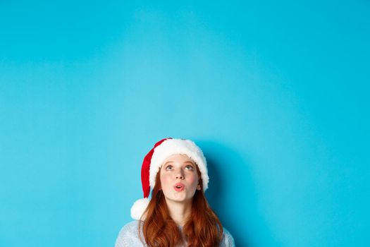 Winter holidays and Christmas eve concept. Head of pretty redhead girl in santa hat, appear from bottom and looking up at logo impressed, seeing promo offer, blue background.