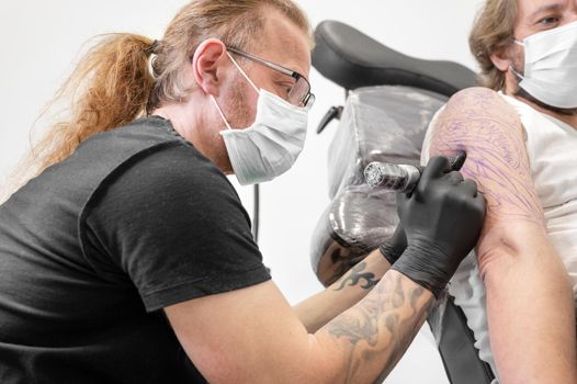 Tattooist with his machine tattooing on the arm with the design drawn and using protective mask against covid 19, concept of art and design during the new normal. High quality photography