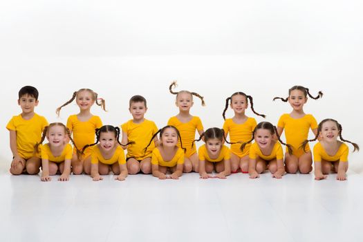 Dancer team sitting on floor in row posing in studio. Group of happy little boys and girls in yellow sportswear posing against white background. Dance school, kids education concept
