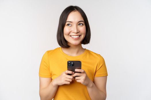 People and technology concept. Smiling asian girl using smartphone, texting on mobile phone, standing against white background.