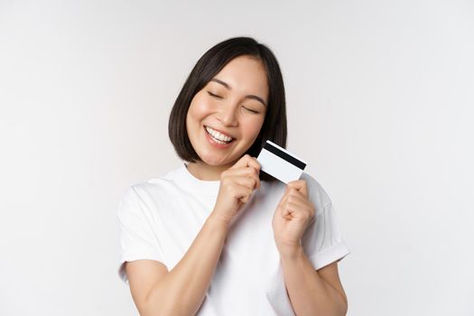 Image of smiling asian woman hugging credit card, buying contactless, standing in white tshirt over white background. Copy space