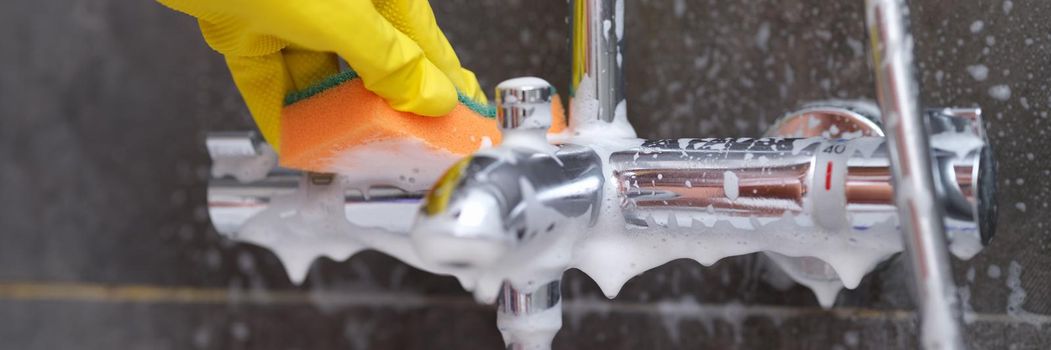 Hand in yellow glove washes chrome-plated mixer with washcloth in bathroom. Plumbing care concept