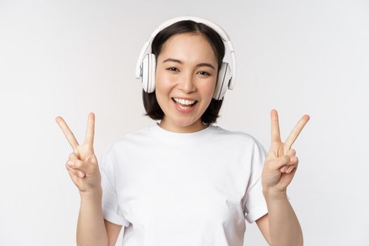 Happy asian woman wearing headphones and smiling, showing peace v-sign, listening music, standing in tshirt over white background.