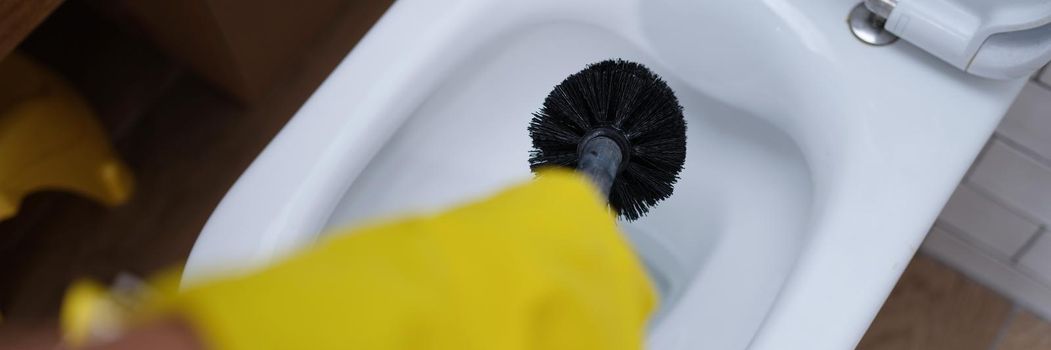 Hand in glove is cleaning toilet bowl with black brush. Cleaning toilet bowl from water and urinary stones concept